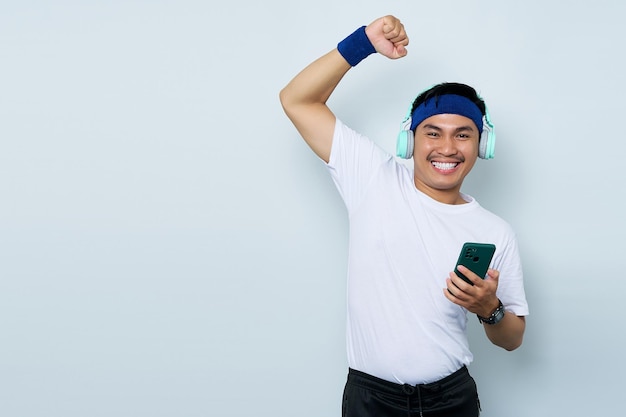 Retrato de alegre jovem desportista asiático na faixa azul e camiseta branca de roupas esportivas enquanto ouve música com fones de ouvido dança ao som de música favorita isolada no fundo branco