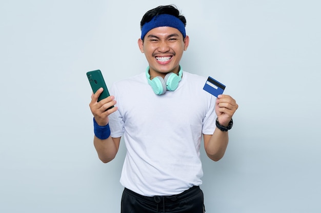 Retrato de alegre jovem desportista asiático na faixa azul e camiseta branca de roupas esportivas enquanto ouve música com fones de ouvido segurando telefone celular e cartão de crédito isolado no fundo branco