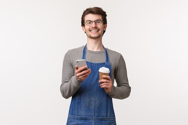 Retrato de alegre jovem empregado do sexo masculino sugerir usar código promocional com o aplicativo do telefone móvel para obter desconto em sua cafeteria, segurando a xícara de take-away e smartphone sorrindo câmera