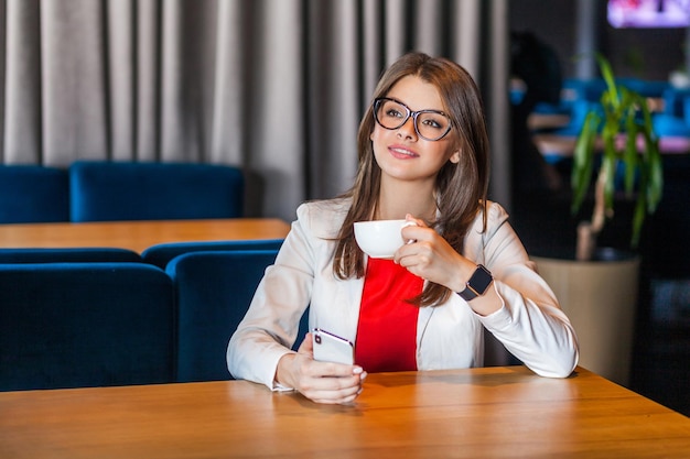 Retrato de alegre linda elegante morena jovem em copos sentado segurando a xícara de café, sorriso, olhando para longe, descansando e curtindo. tiro de estúdio interior, café, fundo de escritório.