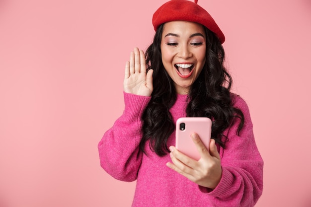 Retrato de alegre linda menina asiática com longos cabelos escuros usando boina, sorrindo e acenando para smartphone isolado em rosa