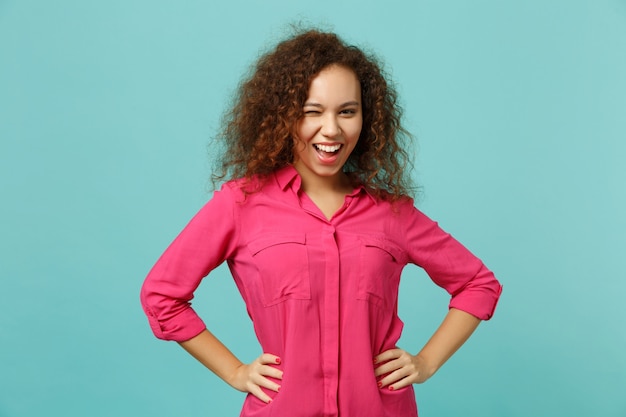 Retrato de alegre menina africana piscando em roupas casuais, em pé com as mãos nos quadris isoladas sobre fundo azul turquesa em estúdio. emoções sinceras de pessoas, conceito de estilo de vida. simule o espaço da cópia.