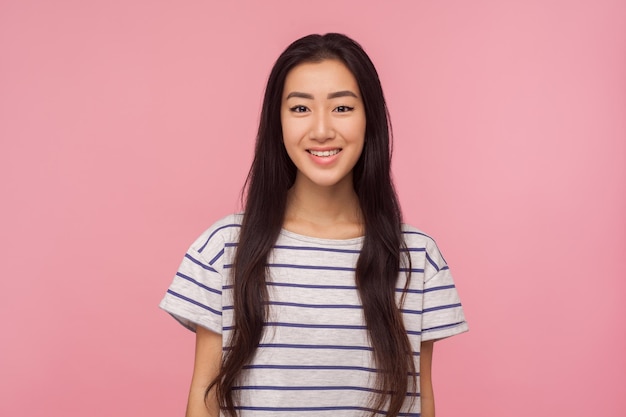 Retrato de alegre menina asiática bonita com longos cabelos morenos em camiseta listrada, olhando para a câmera com sorriso cheio de dentes, aproveitando a vida feliz, bom humor, estúdio interno, tiro isolado no fundo rosa