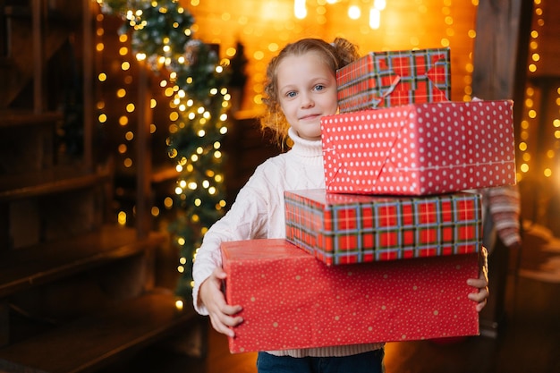 Retrato de alegre menina loira encaracolada segurando muitas caixas de presente com presentes de natal em pé na aconchegante sala de estar com interior festivo