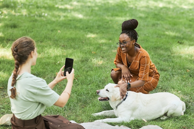 Retrato de alto ângulo de duas mulheres no parque tirando fotos com o cachorro ao ar livre e sorrindo, copie o espaço