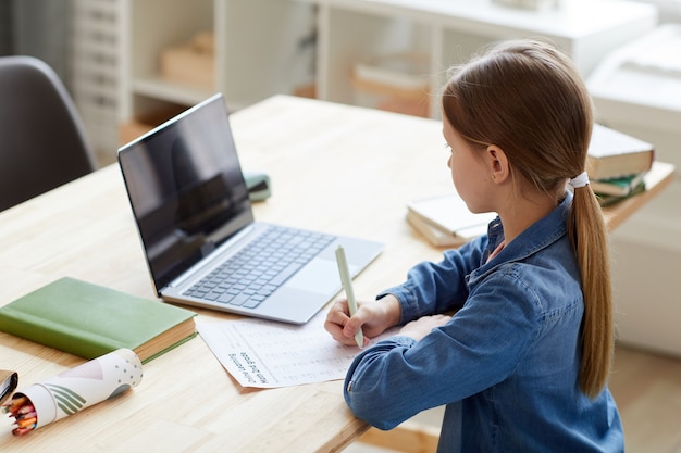 Retrato de alto ângulo de uma menina usando um laptop durante a aula on-line com o tutor ou professor enquanto estudava em casa, sentado na mesa em um interior aconchegante, copie o espaço