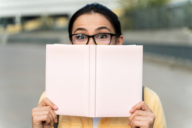 Foto retrato de aluna feliz, cobrindo o rosto com o livro e olhando para a câmera. mulher morena se divertindo e brincando