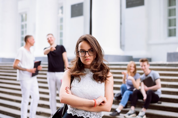 Retrato de aluna infeliz e triste ao ser intimidado por seus colegas de grupo. Alunos fazendo brincadeiras, zombando e zombando de seus colegas de classe nas escadas da faculdade.