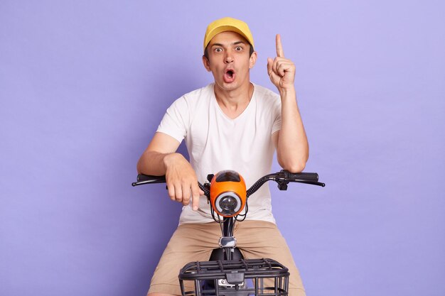 Retrato de animado entregador moreno de takeaway em bicicleta elétrica posando isolado sobre fundo roxo, apontando o dedo para cima tendo uma excelente ideia olhando para a câmera com a boca aberta