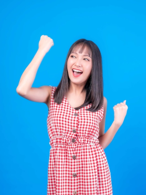 Retrato de animado linda mulher asiática linda garota de vestido vermelho com estilo de cabelo franja levantando a mão no ar rindo alegremente gesticulando para o vencedor sorrindo olhar câmera isolada fundo azul