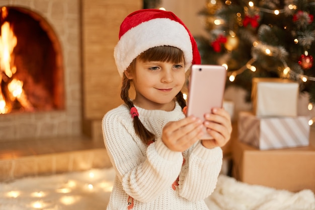 Retrato de ano novo da menina bonita contra pela árvore de Natal e lareira, criança segurando o telefone celular, tem chamada de vídeo e parabéns, alguém, usando chapéu vermelho festivo.