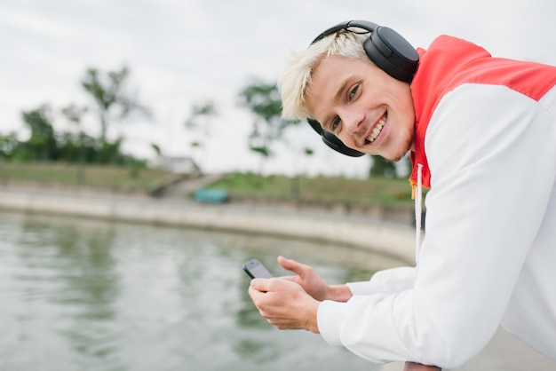 Retrato de atleta de homem sorridente caucasiano atraente relaxar após treino de mensagens ao ar livre através de redes sociais no telefone inteligente e usando fones de ouvido para ouvir música Conceito de estilo de vida saudável