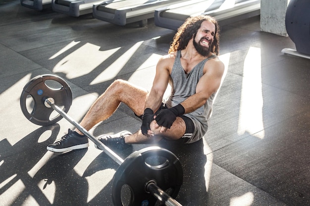 Retrato de atleta jovem adulto chateado com cabelo longo encaracolado malhando na academia sentado no chão e tem forte problema de machucado com lesão no joelho Agarrando a perna com a mão e gritando Indoor