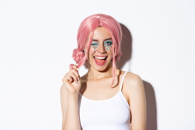 Retrato de atrevida garota atraente na peruca rosa, vestindo roupa para festa de halloween, sorrindo e piscando para a câmera, em pé sobre um fundo branco.