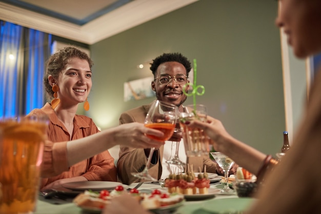 Retrato de baixo ângulo de elegante casal de mestiços jantando com amigos dentro de casa e batendo os copos na mesa festiva, copie o espaço