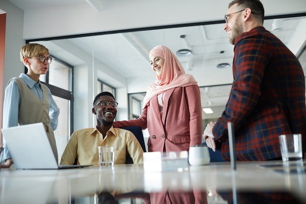 Retrato de baixo ângulo de uma alegre equipe de negócios multiétnica em pé ao lado da mesa de reunião na sala de conferências, com foco na mulher muçulmana com lenço na cabeça ao lado de um colega afro-americano