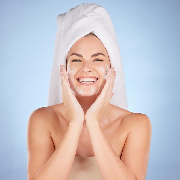 Retrato de banho e mulher lavando o rosto com sabão no fundo azul do estúdio e cosméticos de limpeza Modelo feminino feliz skincare facial e espuma para dermatologia de brilho saudável e beleza hidratante