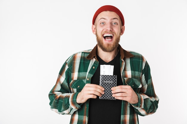 Retrato de barbudo com chapéu e camisa xadrez, segurando o passaporte e as passagens, em pé, isolado sobre um fundo branco