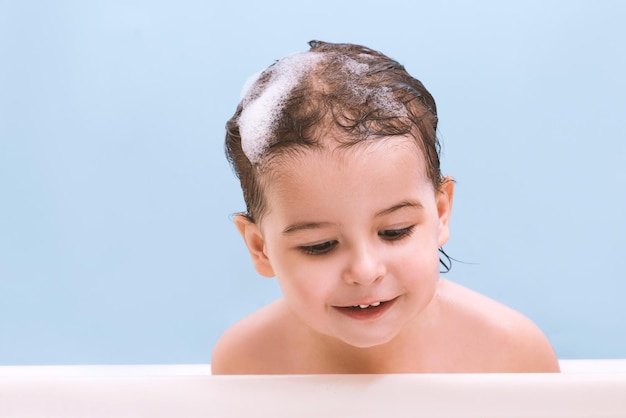 Retrato de bebê bebê fofo tomando banho brincando com bolhas de espuma Criança em uma banheira olhando para baixo Criança no banheiro em fundo azul Banho infantil Higiene e cuidados de saúde