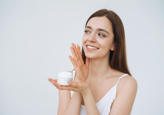 Retrato de beleza de mulher sorridente feliz com cabelos longos escuros colocar creme hidratante nutritivo dia no rosto de pele fresca limpa e mãos no fundo branco isolado