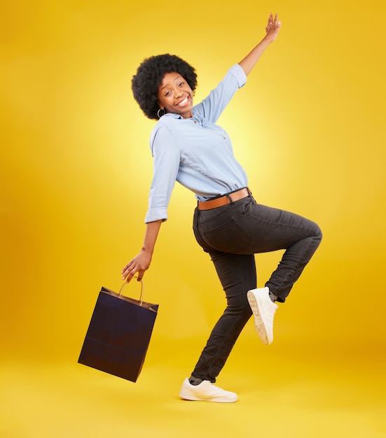 Retrato de bolsa e mulher negra feliz fazendo compras animadas e comemorando a venda no estúdio em fundo amarelo Comprador de rosto e cliente de menina alegre após varejo boutique ou desconto de loja isolado