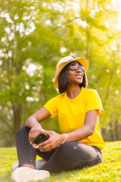 Retrato de brilho de verão de uma jovem turista negra curtindo o pôr do sol em um chapéu de camisa amarela e óculos de sol