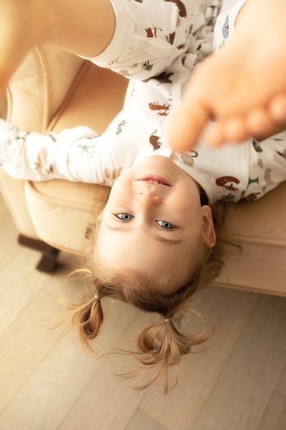 Retrato de cabeça para baixo de uma linda garota ativa emocional adorável criança deitada de cabeça para baixo no biege chear se divertindo dentro de casa