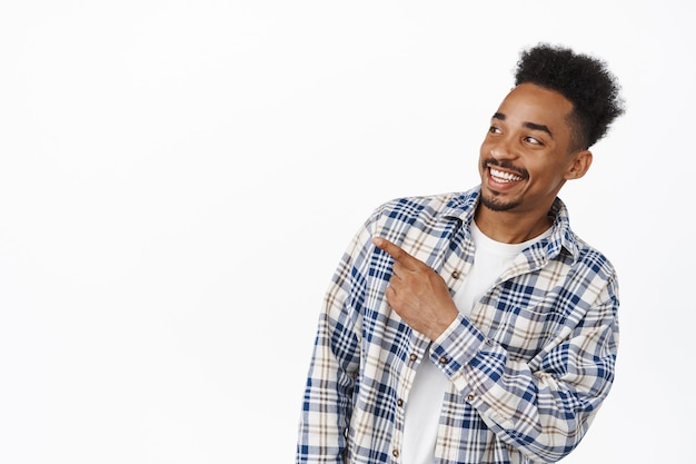 Retrato de cara afro-americano bonito sorrindo feliz, apontando e olhando para a esquerda no banner promo de venda, mostrando o anúncio, em pé em roupas casuais em branco.