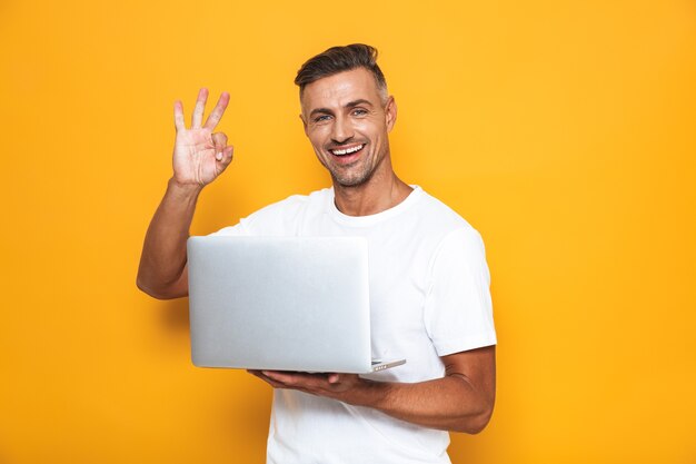 Retrato de cara bonito dos 30 anos em uma camiseta branca segurando e usando laptop prateado isolado em amarelo