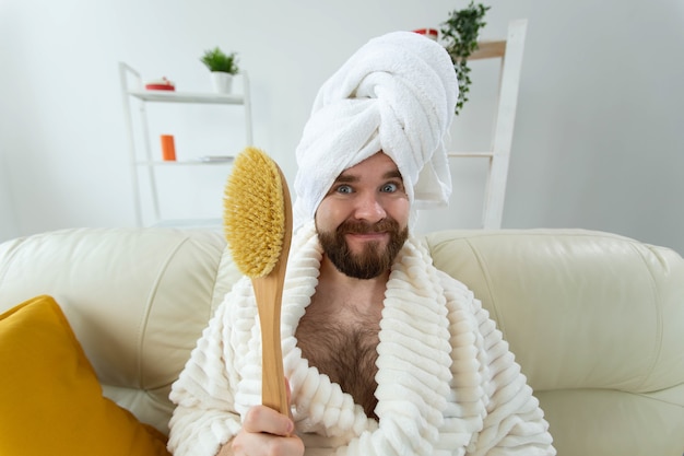 Retrato de cara engraçado usar turbante toalha segurando na escova de massagem de mãos enquanto se senta no sofá. Cuidados com a pele masculina e conceito de spa.