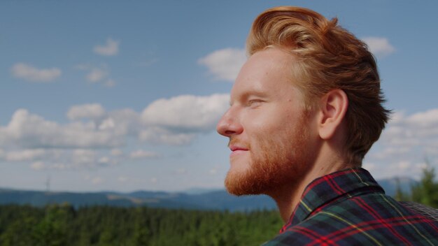 Retrato de cara relaxado, apreciando a paisagem montanhosa no verão Viajante masculino alegre olhando bosques verdes no campo Homem sorridente explorando a natureza Hipster ruiva tendo aventura nas montanhas