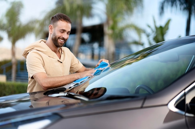 Retrato de carro de limpeza de homem latino sorridente usando pano de microfibra. conceito de serviço de lavagem de carros