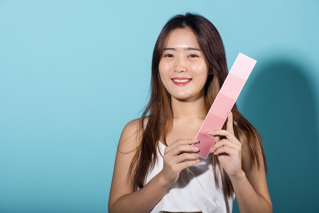 Retrato de cartão de cor de preensão feminina feliz sob proteção solar, rosto de beleza de pele, mulher jovem e bonita asiática sorrindo e segurando papel de escala de cor de pele tiro de estúdio isolado em fundo azul