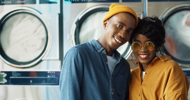 Retrato de casal alegre afro-americano feliz no amor, abraçando e sorrindo para a câmera no serviço de lavanderia. homem novo alegre e mulher que estão em trabalhar máquinas de lavar roupa dentro do washhouse.