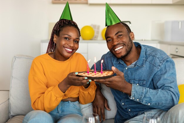 Retrato de casal americano africano em chapéus festivos segurando torta de aniversário com velas acesas e sorrindo para a câmera em casa