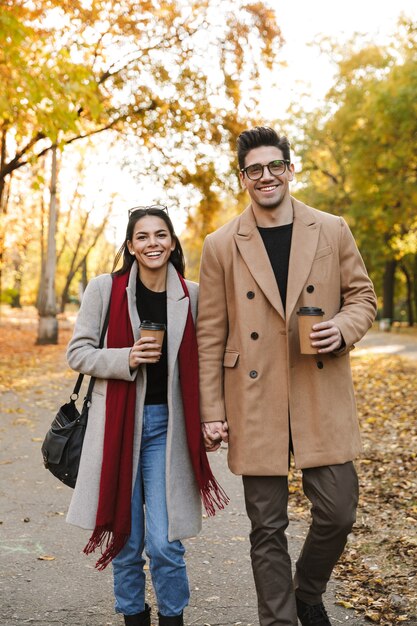 Retrato de casal casual bebendo café para viagem em copos de papel e sorrindo enquanto caminha no parque outono