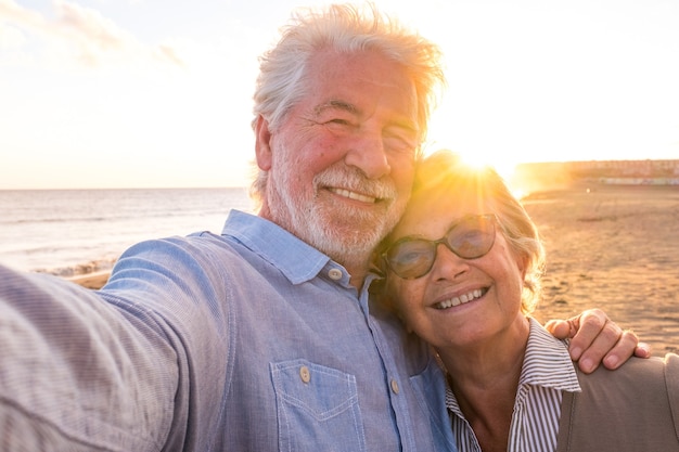 Retrato de casal de pessoas maduras e velhas aproveitando o verão na praia, olhando para a câmera, tirando uma selfie junto com o pôr do sol ao fundo. Dois idosos ativos viajando ao ar livre.
