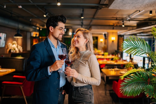 Retrato de casal elegante sorridente apaixonado segurando copos de vinho tinto olhando um para o outro em pé posando em um restaurante chique à noite