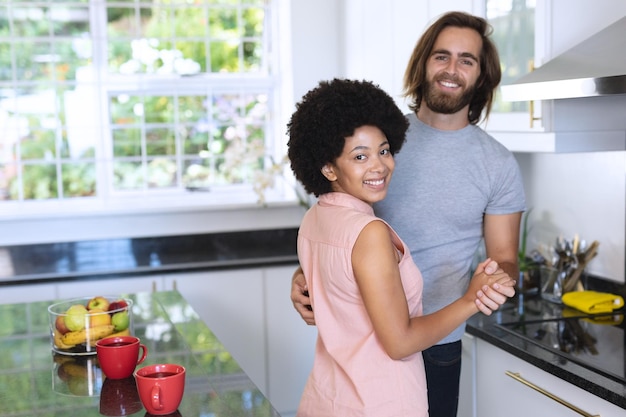 Retrato de casal feliz e diversificado dançando na cozinha e sorrindo