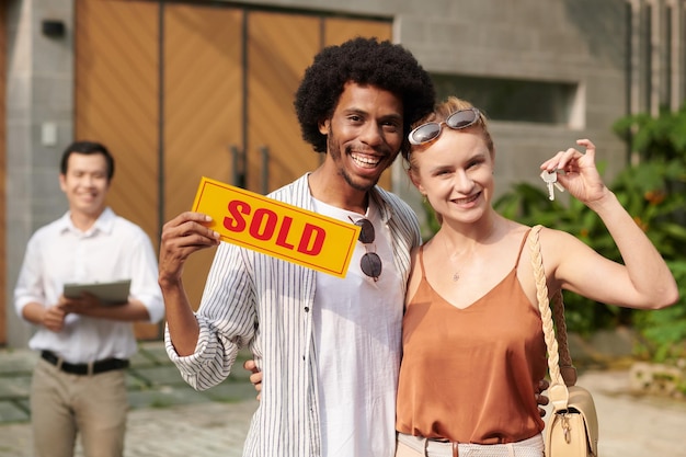 Retrato de casal feliz e diversificado mostrando sinal vendido e chaves da nova casa