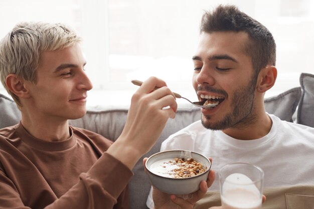 Retrato de casal gay brincalhão tomando café da manhã juntos, foco em jovem sorridente alimentando o namorado