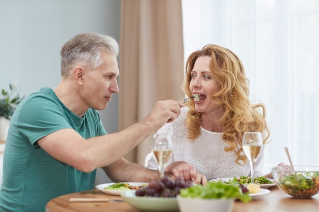 Retrato de casal maduro moderno desfrutando de um jantar juntos em casa, homem dando comida no garfo para a esposa, copie o espaço