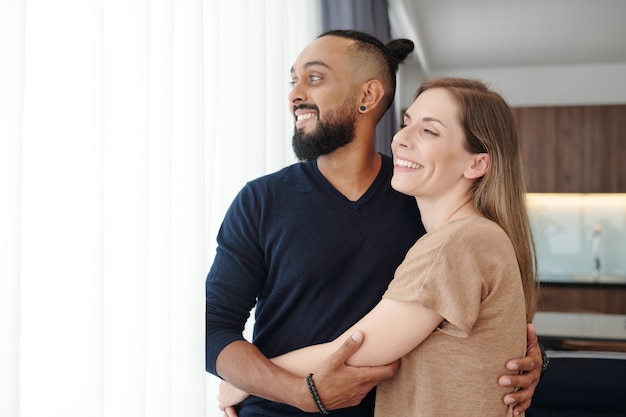 Retrato de casal mestiço se abraçando feliz se abraçando e olhando pela janela