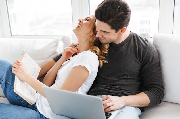Retrato de casal otimista usando laptop e lendo um livro enquanto está sentado no sofá em uma sala iluminada em casa