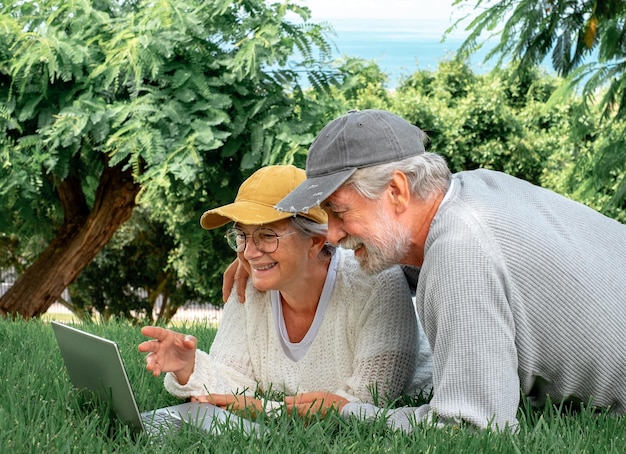 Retrato de casal sorridente sênior deitado na grama no parque público olhando juntos para laptop Casal de idosos modernos aproveitando o tempo livre e a aposentadoria