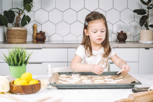 Retrato de chef de menina cuidadosamente sentado na cadeira fazendo biscoitos de massa de forma diferente na pingadeira Placa de madeira de ovos de páscoa amarelos e vaso de plantas perto de criança Utensílio de cozinha interior