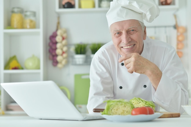 Retrato de chef masculino idoso cozinhando na mesa com laptop