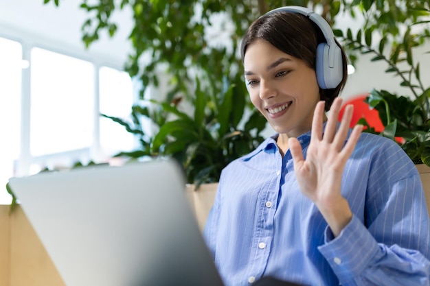 Foto retrato de cintura de uma mulher alegre com fones de ouvido acenando para alguém enquanto olha para o monitor do computador