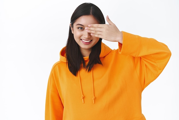 Retrato de cintura linda garota moderna asiática com capuz laranja elegante cobrir um olho com palma e promoção sorridente de produtos de beleza para a pele e maquiagem coreana em pé fundo branco