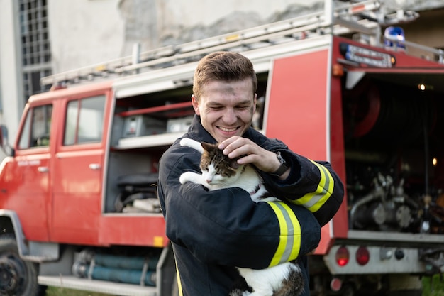 Retrato de close-up de bombeiro heróico em traje de proteção e capacete vermelho guarda gato salvo em seus braços. Bombeiro em operação de combate a incêndio. Foto de alta qualidade